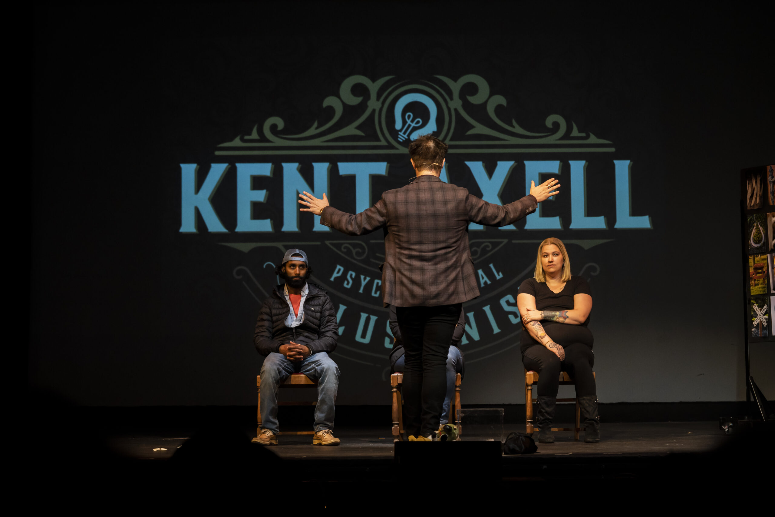 Kent Axell on stage with two volunteers seated beside him